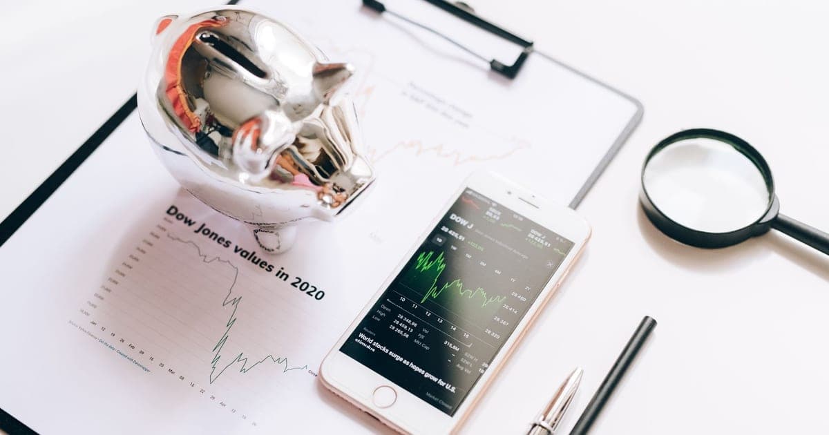 A glass piggy bank beside a printed chart, a phone showing a market graph and a magnifying glass