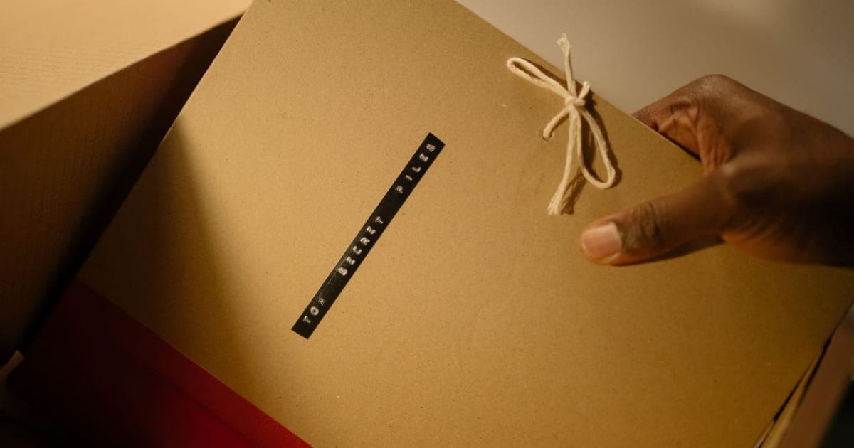 A hand lifting a string-tied cardboard folder labeled top secret files out of a box