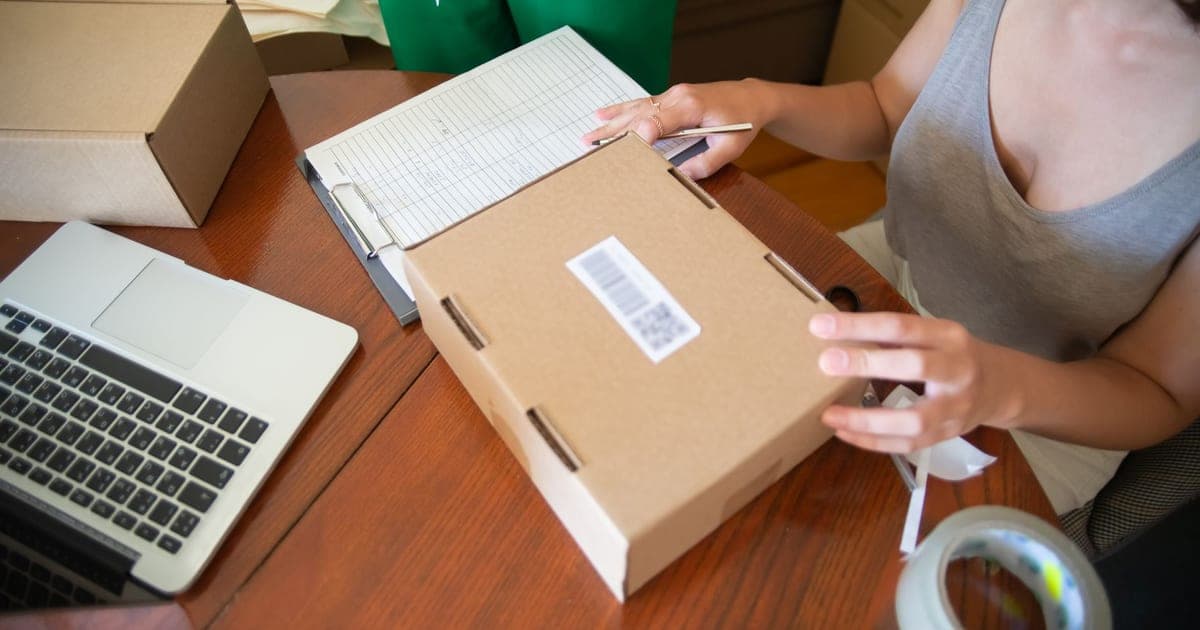 Someone at a desk sealing a cardboard box with a shipping label, an open laptop and an order sheet beside them