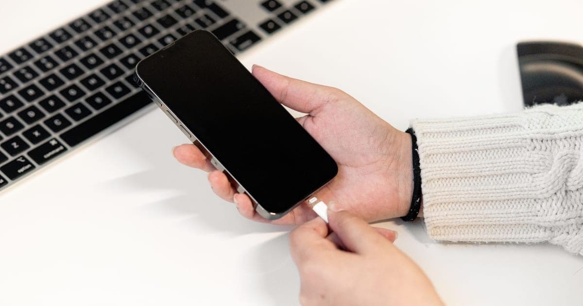 A hand bringing a plain white charging cable to a phone's port over a white desk, the connector indistinguishable from any other