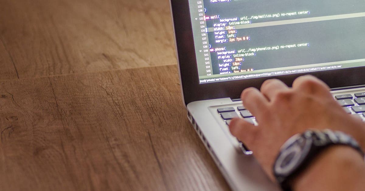 Hands typing on a laptop displaying lines of code at a wooden desk